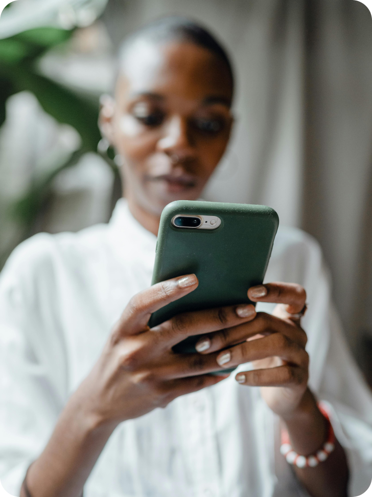 Eine Person hält ein Smartphone in beiden Händen und schaut konzentriert darauf. Sie trägt ein weißes Hemd und hat kurze Haare. Der Hintergrund ist leicht verschwommen, mit einem kleinen Teil einer Pflanze sichtbar. Die Situation deutet darauf hin, dass die Person möglicherweise nach einem Cannabis-Rezept online sucht.